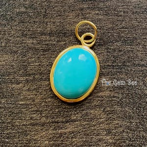 May include: An oval-shaped turquoise pendant set in a gold-toned metal bezel with a circular jump ring at the top. The smooth, bright blue stone is polished and centered within the frame. The text The Gem Bee is visible to the right of the pendant.
