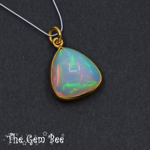 May include: A gold pendant with a pear-shaped opal gemstone. The opal displays iridescent colors of green, orange, and blue. The pendant hangs from a thin white cord. The Gem Bee logo is at the bottom.