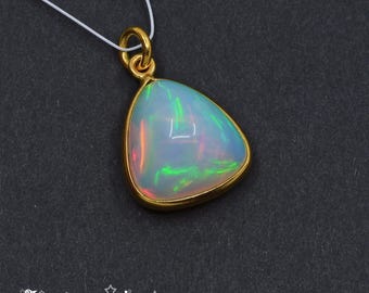 Może przedstawiać: Złoty wisiorek z opalem w kształcie gruszki. Opal wykazuje opalizujące kolory zieleni, pomarańczy i błękitu. Wisiorek zawieszony jest na cienkim, białym sznurku. Logo The Gem Bee znajduje się na dole.