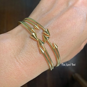 May include: Gold-toned bracelet featuring a series of three parallel bands, adorned with conical spike accents. The bracelet is shown on a wrist, showcasing its design and style. The Gem Bee logo is visible in the bottom right corner.