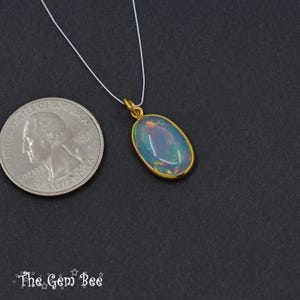 Puede incluir: Colgante de oro con una piedra preciosa de &oacute;palo ovalada, suspendida de un fino cord&oacute;n blanco. El &oacute;palo muestra colores iridiscentes. Una moneda de veinticinco centavos estadounidense est&aacute; al lado del colgante para comparar el tama&ntilde;o. El logo de The Gem Bee est&aacute; abajo.