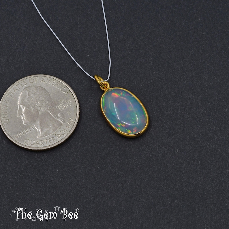 Puede incluir: Colgante de oro con una piedra preciosa de &oacute;palo ovalada, suspendida de un fino cord&oacute;n blanco. El &oacute;palo muestra colores iridiscentes. Una moneda de veinticinco centavos estadounidense est&aacute; al lado del colgante para comparar el tama&ntilde;o. El logo de The Gem Bee est&aacute; abajo.