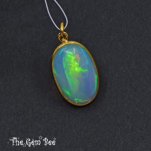 May include: Oval opal pendant with a gold-coloured bezel and bail. The opal displays a play of colour with blue, green, and yellow hues. The pendant hangs from a thin white cord. The Gem Bee logo is in the bottom left corner.