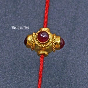 May include: A gold bead with four red gemstones set into the bead. The bead is strung on a red cord.
