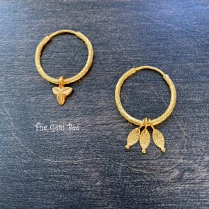 Puede incluir: Un par de pendientes de aro dorados con superficies texturizadas. Un pendiente presenta un dije de tres p&eacute;talos, mientras que el otro muestra tres dijes colgantes en forma de hoja. El texto The Gem Bee es visible en la esquina inferior izquierda.