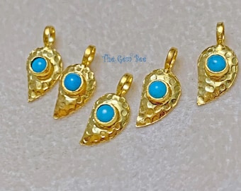 May include: Five gold-toned metal pendants shaped like teardrops or leaves, each featuring a hammered texture and a single round, bright blue stone set in the center. Each pendant includes a small loop at the top. The text The Gem Bee is visible.