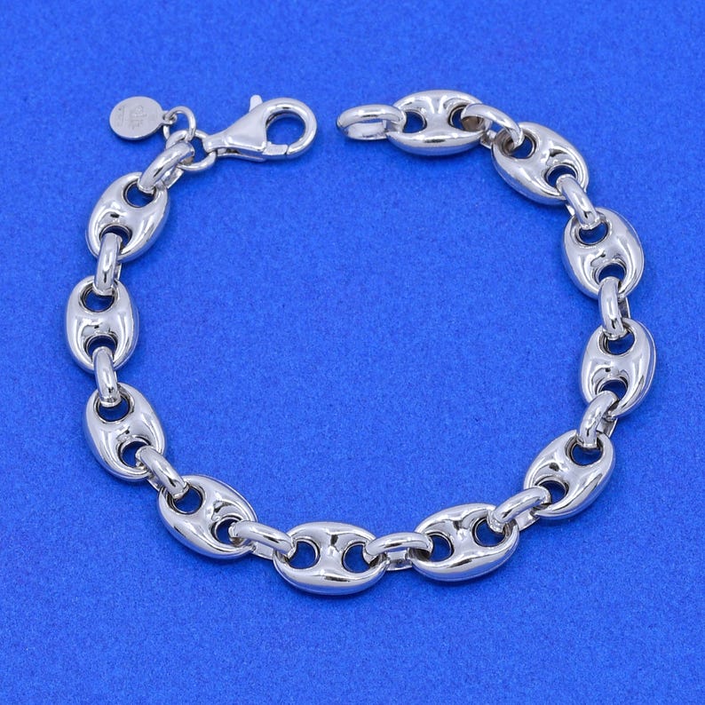 May include: A silver-colored bracelet with a series of oval-shaped links, connected to each other. The bracelet has a lobster clasp closure and a small circular charm. The jewelry is displayed against a blue background.