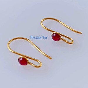 Könnte beinhalten: Ein Paar goldfarbene Ohrhänger, jeweils mit einem kleinen, runden, dunkelroten Cabochon-Stein an der Basis des gebogenen Drahtes. Der Text The Gem Bee ist in Blau zwischen den beiden Ohrringen auf einem schlichten, hellen Hintergrund gedruckt.