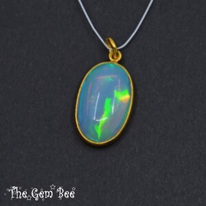 May include: Oval opal pendant with a gold-coloured bezel, suspended from a thin white cord. The opal displays a play of colour with blue, green, and yellow hues. The Gem Bee logo is in the bottom left corner.