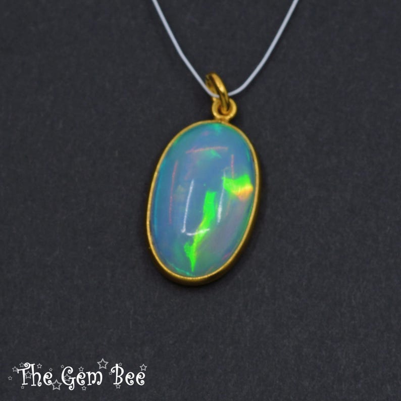 May include: Oval opal pendant with a gold-coloured bezel, suspended from a thin white cord. The opal displays a play of colour with blue, green, and yellow hues. The Gem Bee logo is in the bottom left corner.