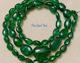 Puede incluir: Un collar de cuentas de color verde esmeralda pulidas y de forma ovalada dispuestas en un patrón circular. Las cuentas varían en tamaño, con piedras más grandes colocadas en la parte inferior. El texto The Gem Bee está impreso en azul.