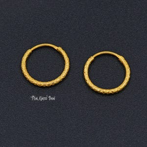 Puede incluir: Un par de pendientes de aro dorados con un patr&oacute;n texturizado y repetitivo en toda la superficie de los aros circulares. Las piezas de joyer&iacute;a se muestran sobre un fondo oscuro y granulado con el texto The Gem Bee visible debajo.