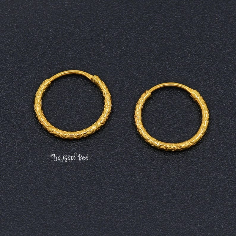 Puede incluir: Un par de pendientes de aro dorados con un patr&oacute;n texturizado y repetitivo en toda la superficie de los aros circulares. Las piezas de joyer&iacute;a se muestran sobre un fondo oscuro y granulado con el texto The Gem Bee visible debajo.