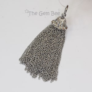 Puede incluir: Un colgante de borla de cadena plateada con una parte superior de cristal transparente. La borla está hecha de muchas pequeñas cadenas plateadas.