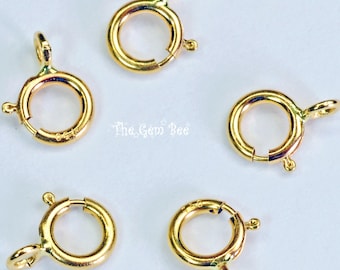 May include: Five gold-toned circular spring ring clasps for jewelry making, each featuring a small attached loop and a spring-loaded mechanism. The text The Gem Bee is printed in the center of the arrangement of these metallic jewelry findings.