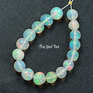 May include: A strand of white opal beads with iridescent rainbow flashes. The beads are round and smooth, and they are strung on a clear thread. The Gem Bee is printed on a white label in the center of the image.