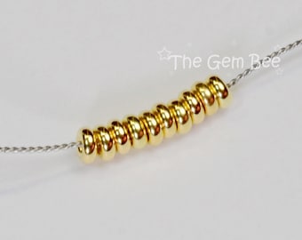 May include: A silver-toned wire necklace featuring a row of ten smooth, polished gold-colored round beads. The text "The Gem Bee" is visible in the upper right corner. The beads are arranged horizontally along the thin, twisted metallic wire strand.