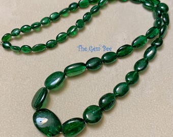 Puede incluir: Un collar con cuentas ovaladas de color verde esmeralda, pulidas y translúcidas, dispuestas en un patrón graduado con las cuentas más grandes en el centro. El texto "The Gem Bee" está impreso en azul cerca del centro del collar.