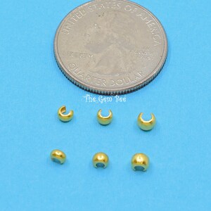 May include: Six gold-coloured crimp beads, each about the size of a UK 20 pence coin, are arranged on a blue surface. The beads are used for jewellery making.