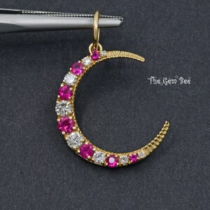 May include: A gold-coloured crescent moon pendant, studded with alternating pink and clear gemstones. The pendant is held by tweezers. The text "The Gem Bee" is visible.