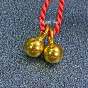 May include: Two polished gold-tone spherical charms suspended from a twisted red cord. The text "The Gem Bee" is printed in white above the charms. The charms feature smooth, reflective surfaces and small loops at the top for attachment.