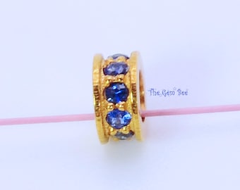 May include: A gold-toned cylindrical bead featuring a central row of round, deep blue faceted stones. The bead has raised edges and is threaded onto a thin pink string. The text The Gem Bee is printed in blue to the right of the bead.