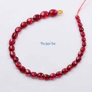 Könnte beinhalten: Eine Strähne ovaler, tief roter Edelsteinperlen. Die Perlen sind auf einem dünnen roten Faden aufgefädelt und bereit, zu einem Armband oder einer Halskette verarbeitet zu werden.