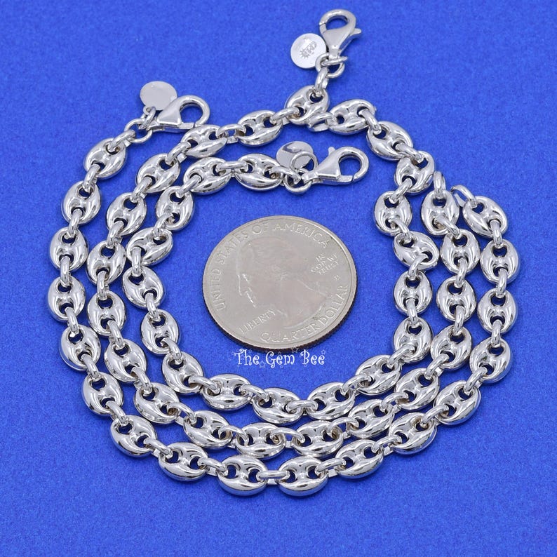 May include: A close-up shot of a silver-colored chain necklace with a unique puffed mariner link design. The necklace is displayed on a blue surface next to a quarter coin for size reference. The Gem Bee logo is visible.