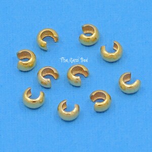 May include: Ten gold-coloured crimp beads, also known as crimp covers, are scattered on a blue background. These beads are used to secure jewellery findings and create a professional finish.