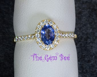 Op de afbeelding: Een gouden ring met een centrale ovaal geslepen blauwe edelsteen omringd door een halo van kleine witte steentjes. De ringband is ook bezet met kleine witte steentjes aan de zijkanten. De tekst The Gem Bee staat in het paars afgedrukt.