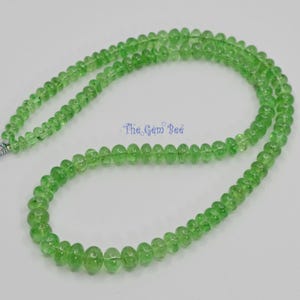 May include: A strand of translucent, light green, rondelle-shaped beads arranged in a circular loop. The beads have a smooth, polished finish and are strung together on a thin cord. The text The Gem Bee is printed in blue in the center.