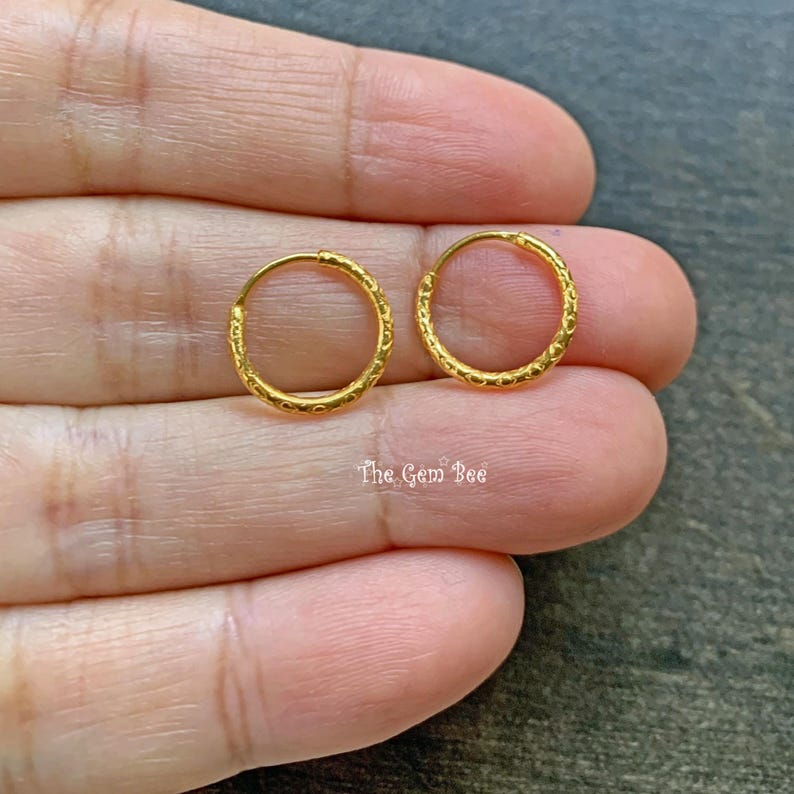 Puede incluir: Un par de peque&ntilde;os pendientes de aro dorados y circulares que presentan un delicado patr&oacute;n texturizado en la superficie exterior. Los aros son finos y redondos, con un acabado en oro amarillo brillante. El texto The Gem Bee es visible.