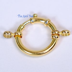 May include: A gold-toned circular spring ring clasp featuring a smooth, polished finish. The jewelry component includes two small, fixed loops extending from opposite sides of the ring and a small spring-loaded mechanism. Text reads The Gem Bee.