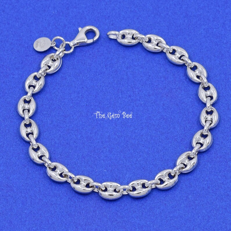 May include: A silver-colored bracelet with a bean-shaped link design. The bracelet has a lobster clasp closure and a small, round charm. The background is a solid blue color. The text "The Gem Bee" is visible.