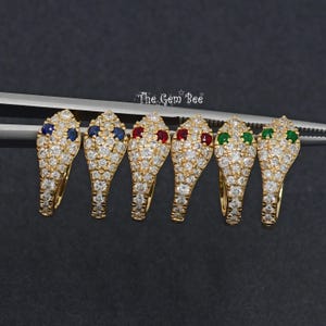 May include: Six gold-colored snake-shaped earrings with diamond accents and colored gemstone eyes. The eyes are blue, red, and green. The earrings are held by a silver tweezer against a dark background. The Gem Bee logo is in the upper right.