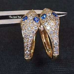 May include: A pair of gold-colored snake-shaped earrings. The earrings are encrusted with small, clear gemstones and feature blue gemstones for the eyes. The earrings are on a dark background.