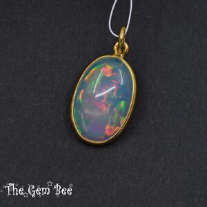 Puede incluir: Colgante de &oacute;palo ovalado con bisel y fianza dorados. El &oacute;palo muestra colores iridiscentes de azul, verde y rojo. El colgante cuelga de un fino cord&oacute;n blanco sobre un fondo oscuro. El logotipo de The Gem Bee est&aacute; en la esquina inferior izquierda.