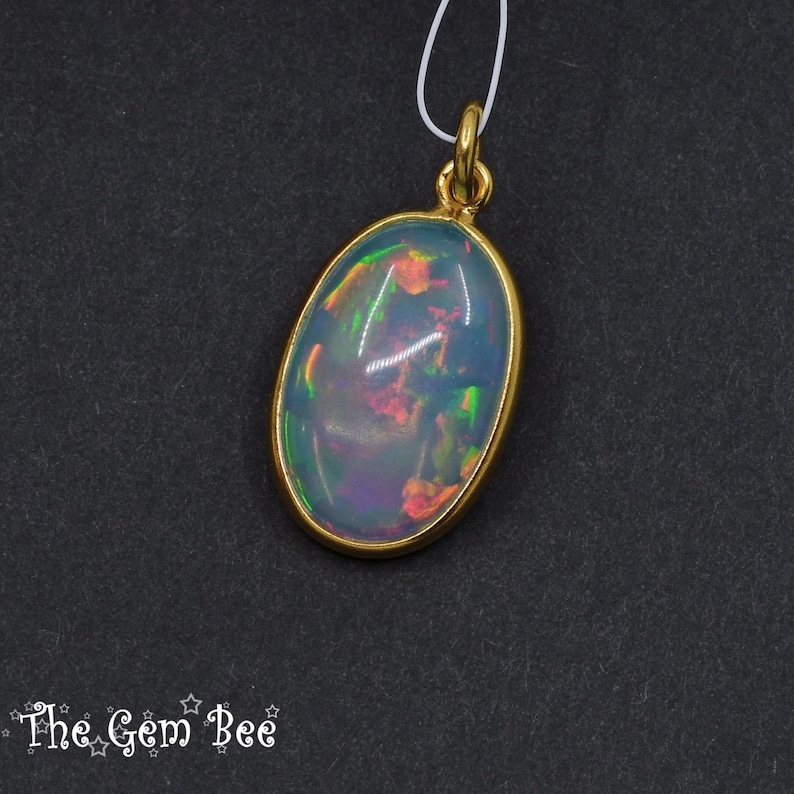 Puede incluir: Colgante de &oacute;palo ovalado con bisel y fianza dorados. El &oacute;palo muestra colores iridiscentes de azul, verde y rojo. El colgante cuelga de un fino cord&oacute;n blanco sobre un fondo oscuro. El logotipo de The Gem Bee est&aacute; en la esquina inferior izquierda.