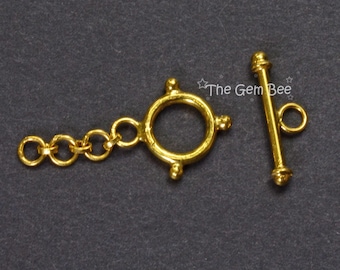 May include: A gold-toned toggle clasp for jewelry making, featuring a circular ring with four small decorative spheres and a matching T-bar component. A short chain of three jump rings is attached to the circular ring. Text reads The Gem Bee.