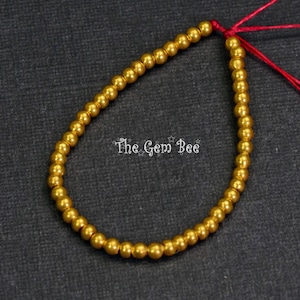 May include: A strand of small, round, gold-colored beads strung on a red cord. The beads are evenly spaced and appear to be made of a metallic material. The text "The Gem Bee" is visible in the image.
