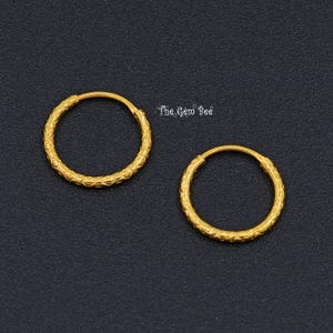 Puede incluir: Un par de pendientes de aro dorados con un patr&oacute;n texturizado y repetitivo en la superficie de los aros circulares. El texto The Gem Bee es visible en letras peque&ntilde;as, blancas y estilizadas situadas entre los dos aros dorados.