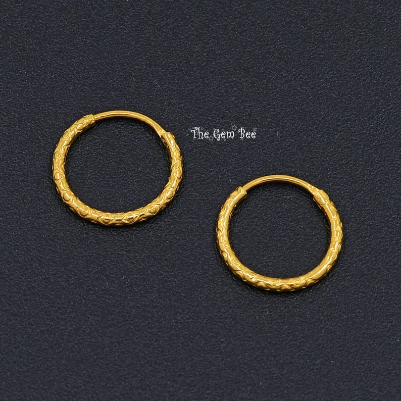 Puede incluir: Un par de pendientes de aro dorados con un patr&oacute;n texturizado y repetitivo en la superficie de los aros circulares. El texto The Gem Bee es visible en letras peque&ntilde;as, blancas y estilizadas situadas entre los dos aros dorados.
