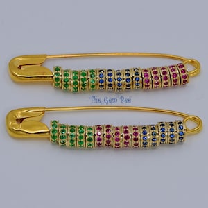 May include: Two gold-colored safety pins with rows of colorful gemstones. Each pin features green, blue, and red gemstones. The text "The Gem Bee" is visible on the image.