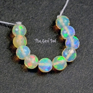 May include: A strand of faceted opal beads, showcasing a play of color with flashes of green, blue, and orange. The beads are round and strung on a white thread. The Gem Bee is printed on the image.