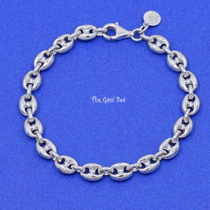 May include: A silver-colored bracelet with a puffed mariner chain design. The bracelet features a lobster clasp closure and a small charm. The Gem Bee text is visible on the blue background.