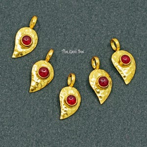 May include: Five gold-colored leaf-shaped pendants with hammered textures, each featuring a round, red gemstone. The pendants are arranged on a dark gray surface. The text "The Gem Bee" is visible.