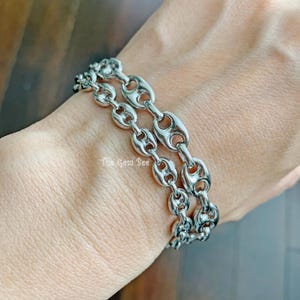 May include: A silver-toned bracelet with a double-strand design. The bracelet features a series of oval-shaped links, creating a bold and stylish accessory. The links are connected by smaller, rounded connectors. The Gem Bee is printed on the image.