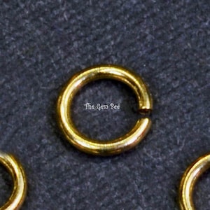 May include: A gold-colored metal jump ring with a circular shape and a small opening. The surface of the ring is smooth and reflective. The text "The Gem Bee" is printed in white in the center of the ring, with small star icons included.