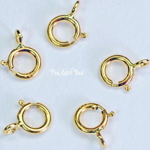 May include: Five gold-toned circular spring ring clasps for jewelry making, each featuring a small attached loop and a spring-loaded mechanism. The text The Gem Bee is printed in the center of the arrangement of these metallic jewelry findings.