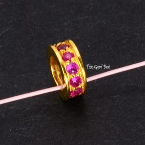 May include: A gold-toned cylindrical bead featuring a central band of vibrant, round-cut pink gemstones. The bead is threaded onto a thin, light pink string. The text The Gem Bee is visible in white lettering to the right of the bead.